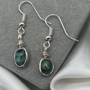 May include: A pair of silver earrings featuring green oval-shaped stones. The stones are set in silver wire, creating a simple and elegant design.