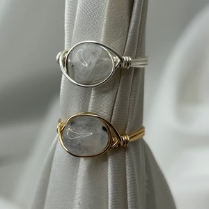 Puede incluir: Dos anillos envueltos en alambre con piedras ovaladas transparentes. El anillo superior está envuelto en alambre plateado, y el anillo inferior está envuelto en alambre dorado.