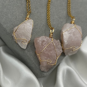 Può includere: Tre collane con pendenti in quarzo rosa grezzo, ciascuno avvolto in filo dorato e sospeso a una catena dorata. Le pietre rosa pallido sono esposte su una superficie grigia con raso bianco.