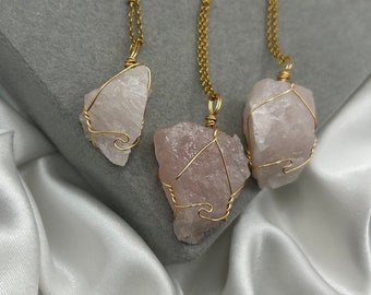 May include: Three rose quartz necklaces with gold-colored wire wrapping and chains. The raw, light pink stones are irregularly shaped. The necklaces are displayed on a gray surface with a white satin fabric.