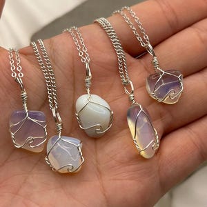 May include: Five gemstone necklaces with silver chains. The gemstones are various shapes and colours, including purple, white, and opalescent. Each stone is wrapped in silver wire. The necklaces are displayed on a hand.