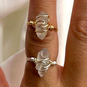 Könnte beinhalten: Zwei Ringe mit klaren Quarz-Kristallsteinen. Ein Ring ist goldfarben, der andere silberfarben. Die Kristalle sind mit Draht in einem wellenartigen Design umwickelt. Die Ringe befinden sich an den Fingern einer Person.