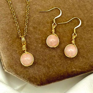 May include: A gold-coloured jewellery set comprising a necklace and earrings. The necklace has a fine chain with a pendant, and the earrings are drop style. Each piece features a light pink, round gemstone, wrapped in gold wire.