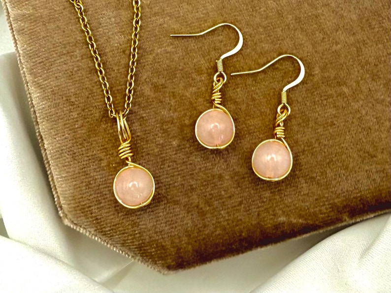 May include: A gold-coloured jewellery set comprising a necklace and earrings. The necklace has a fine chain with a pendant, and the earrings are drop style. Each piece features a light pink, round gemstone, wrapped in gold wire.