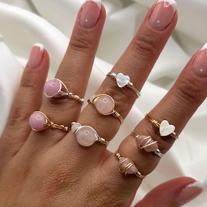 May include: A hand wearing a set of seven rings. The rings are made of gold wire and feature pink and white stones. Some of the stones are heart-shaped.