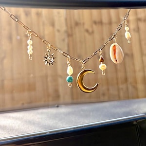 May include: Gold chain necklace featuring a crescent moon, sunburst, pearls, turquoise beads, a seashell, and other decorative beads. The necklace is set against a blurred wooden backdrop.