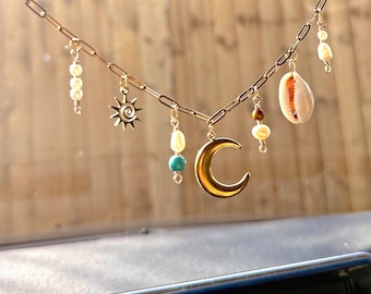 Puede incluir: Collar de cadena dorada con varios dijes, incluyendo una media luna, un sol, perlas, cuentas turquesas, una concha y otras cuentas decorativas. El collar se muestra sobre un fondo de madera borroso.