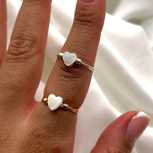 Puede incluir: Dos anillos hechos a mano con dijes en forma de corazón. Un anillo tiene una banda de alambre plateado, el otro una banda de alambre dorado. Los dijes de corazón son blancos. Los anillos se muestran en un dedo sobre un fondo blanco.