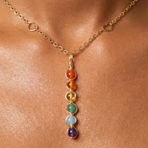 May include: A gold-toned necklace featuring a delicate chain with small heart-shaped links. The pendant consists of six round, multi-coloured gemstone beads arranged vertically and secured with gold-toned wire wrapping in a linear design.