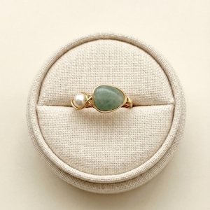 May include: A gold-coloured wire-wrapped ring featuring a light green, irregularly shaped stone and a small white pearl. The ring is displayed on a beige, circular jewellery holder.