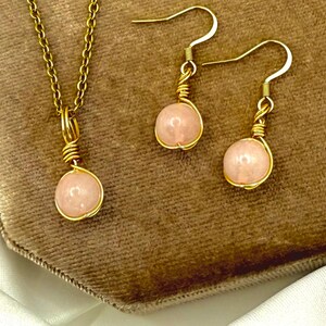 May include: A gold-coloured jewellery set comprising a pendant necklace and a pair of earrings. Each piece features a light pink, round stone encased in gold wire. The necklace has a fine chain, and the earrings have hook fastenings.
