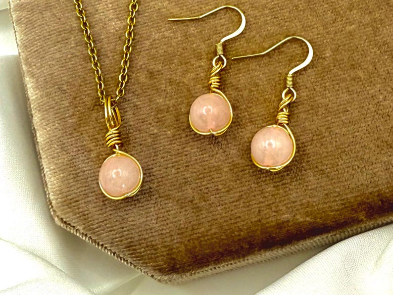 May include: A gold-coloured jewellery set comprising a pendant necklace and a pair of earrings. Each piece features a light pink, round stone encased in gold wire. The necklace has a fine chain, and the earrings have hook fastenings.