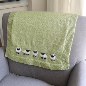 May include: A pale green knitted blanket featuring five embroidered sheep along the lower edge. The blanket is draped over the back of a grey armchair. The sheep have white bodies and black faces and legs.