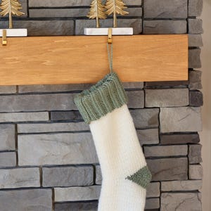 May include: A knitted Christmas stocking featuring a white body with a sage green ribbed cuff, heel, and toe. The stocking hangs from a gold-toned metal tree-shaped hook with a white marble base, positioned on a wooden fireplace mantel.