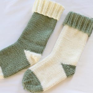 May include: Two hand-knitted stockings featuring a color-blocked design. One stocking is primarily sage green with a cream cuff, heel, and toe. The second stocking is primarily cream with a sage green cuff, heel, and toe. Both have ribbed cuffs.