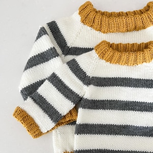 May include: A white and grey striped sweater with a mustard yellow ribbed trim around the neck and cuffs.