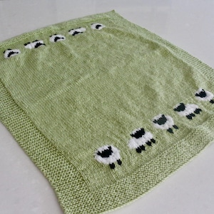 Pu&ograve; includere: Una coperta lavorata a maglia verde chiaro con bordo. La coperta presenta un motivo di pecore bianche e nere sparse sulla superficie. Le pecore sono rivolte in direzioni diverse.