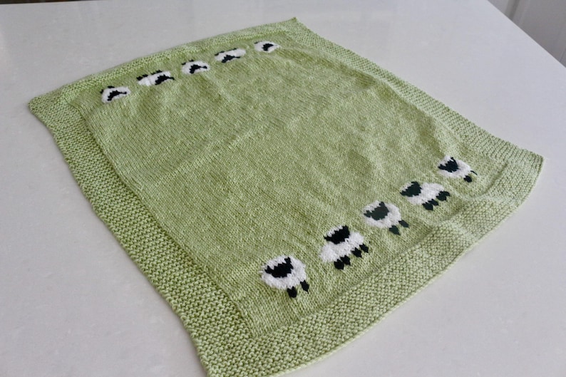 Pu&ograve; includere: Una coperta lavorata a maglia verde chiaro con bordo. La coperta presenta un motivo di pecore bianche e nere sparse sulla superficie. Le pecore sono rivolte in direzioni diverse.