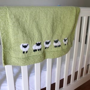 Pu&ograve; includere: Una coperta per neonati lavorata a maglia verde chiaro con un motivo a pecore. La coperta &egrave; drappeggiata sul lato di un lettino bianco. Le pecore sono bianche con facce e zampe nere. La coperta ha un bordo.