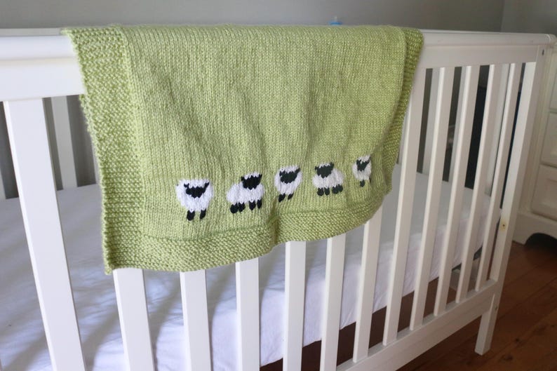Pu&ograve; includere: Una coperta per neonati lavorata a maglia verde chiaro con un motivo a pecore. La coperta &egrave; drappeggiata sul lato di un lettino bianco. Le pecore sono bianche con facce e zampe nere. La coperta ha un bordo.