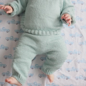 May include: A pale green knitted baby outfit, comprising a jumper and trousers. The outfit is made of soft yarn and features a ribbed waistband and cuffs. The baby is lying on a white sheet with a blue truck pattern.