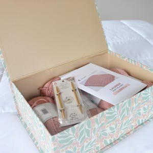 May include: Open box with a floral design containing knitting supplies. The box holds pink yarn, knitting needles, and a pattern for a baby blanket. The needles are US size 8 (5 mm) and 36 inches (90 cm) long.