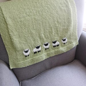 Pu&ograve; includere: Una coperta lavorata a maglia verde chiaro con una fila di cinque applicazioni di pecore lungo il bordo inferiore. Le pecore sono bianche con facce e zampe nere. La coperta &egrave; drappeggiata su una poltrona grigia.