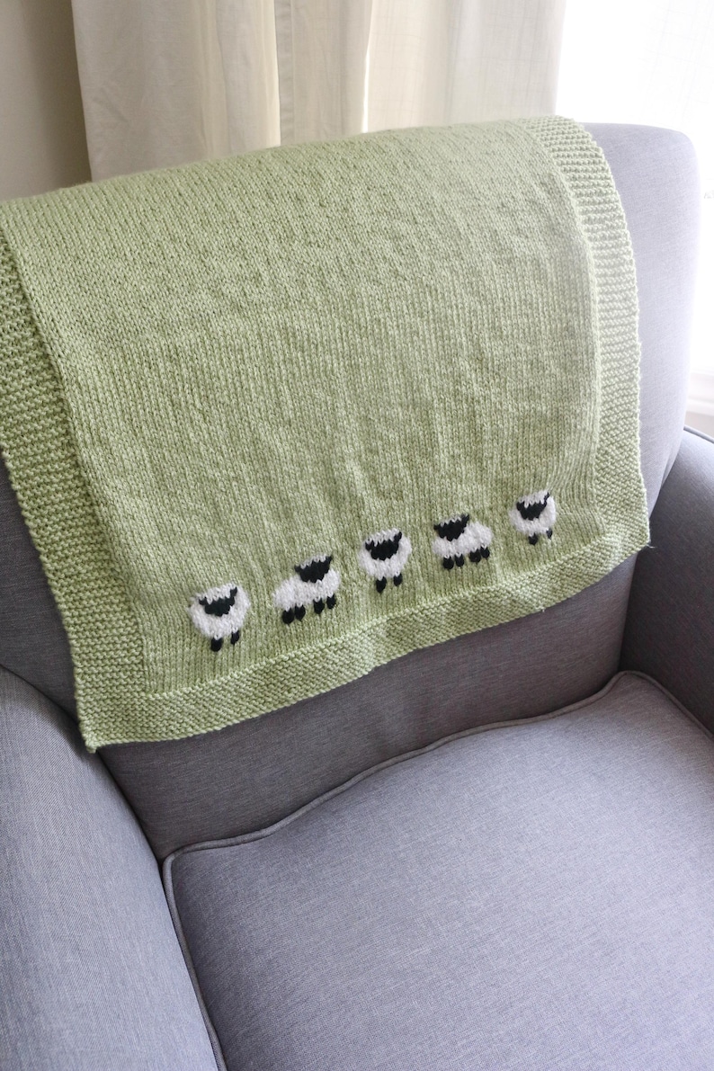 Pu&ograve; includere: Una coperta lavorata a maglia verde chiaro con una fila di cinque applicazioni di pecore lungo il bordo inferiore. Le pecore sono bianche con facce e zampe nere. La coperta &egrave; drappeggiata su una poltrona grigia.