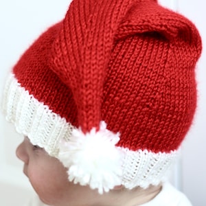 May include: A red and white knitted Santa hat with a white pom-pom on top. The hat is being worn by a baby.