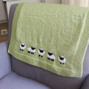 Pu&ograve; includere: Una coperta lavorata a maglia verde chiaro con una fila di cinque pecore ricamate. La coperta &egrave; piegata su una poltrona grigia. Le pecore sono bianche con facce e zampe nere. La coperta ha un bordo a coste.