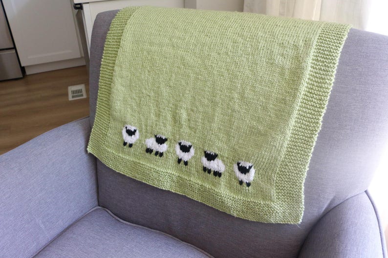 Pu&ograve; includere: Una coperta lavorata a maglia verde chiaro con una fila di cinque pecore ricamate. La coperta &egrave; piegata su una poltrona grigia. Le pecore sono bianche con facce e zampe nere. La coperta ha un bordo a coste.