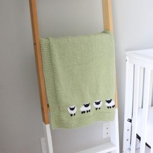 Pu&ograve; includere: Una coperta per neonati lavorata a maglia verde chiaro con un motivo a pecore. La coperta &egrave; drappeggiata su una scala di legno e bianca. Le pecore sono bianche con facce e zampe nere. Sullo sfondo c'&egrave; una culla bianca.