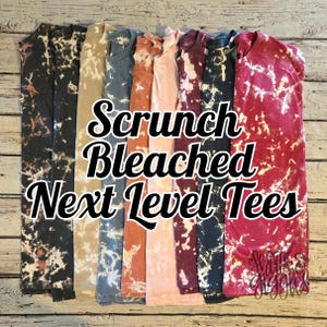 May include: A collection of short-sleeve t-shirts featuring a bleached, mottled pattern in various colors including black, tan, blue, orange, pink, maroon, and red. The text "Scrunch Bleached Next Level Tees" is overlaid in the center of the image.