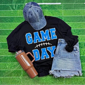 May include: A blue acid-wash baseball cap, a black long-sleeve shirt with blue text reading GAME DAY and a football graphic, a pair of light blue denim shorts with a frayed hem, and a brown tumbler with a white football lace design.