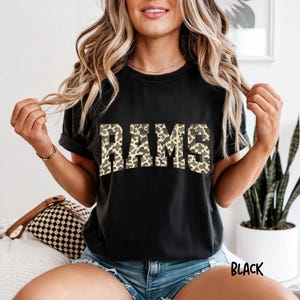 Könnte beinhalten: Ein schwarzes Kurzarm-T-Shirt mit dem Wort RAMS, das in einer Blockschrift auf der Brust gedruckt ist. Die Buchstaben haben ein beiges und braunes Leopardenmuster. Das T-Shirt hat einen Rundhalsausschnitt und eine Standardpassform in Schwarz.