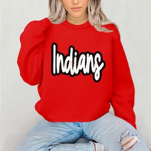 Könnte beinhalten: Ein leuchtend rotes Langarm-Sweatshirt mit Rundhalsausschnitt, das mittig auf der Brust das Wort Indians in einer weißen, abgenutzten Schreibschrift mit dickem schwarzem Rand zeigt. Das Kleidungsstück hat lange Ärmel und einen gerippten Kragen.