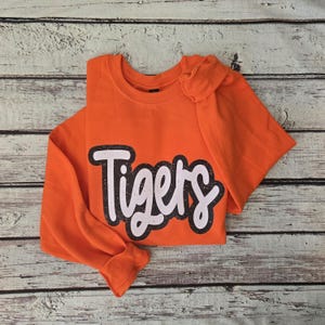 May include: A bright orange long-sleeve crewneck sweatshirt featuring the word Tigers printed in a large, white, cursive font with a distressed black outline. The garment has a classic round neckline and long sleeves with ribbed cuffs.