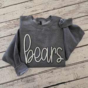 May include: A dark gray long-sleeve crewneck sweatshirt featuring the word bears in large, white, cursive-style lettering across the front. The garment has a ribbed collar, cuffs, and hem, with a soft, fleece-like texture visible on the fabric.