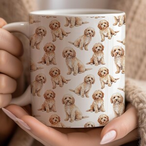 Könnte beinhalten: Weiße Keramiktasse mit einem sich wiederholenden Muster aus Aquarellillustrationen von kleinen, hellbraunen Hunden. Die Hunde sind in verschiedenen Posen dargestellt. Die Tasse hat einen weißen Henkel und wird von einer Person gehalten.