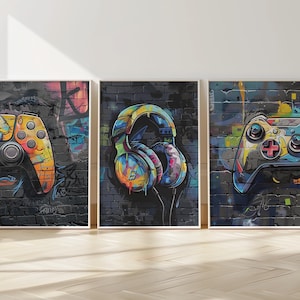Op de afbeelding: Drie ingelijste kunstdrukken met graffiti-achtige illustraties van gamingaccessoires. De prints tonen een gele en oranje gamecontroller, een kleurrijke koptelefoon en een tweede controller. Het kunstwerk is geplaatst tegen een zwarte bakstenen muur.
