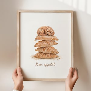 Może przedstawiać: Oprawiony wydruk akwarelowy przedstawiający małego, brązowego psa spoczywającego na stosie rogalików. Słowa "Bon appétit" są napisane kursywą na dole. Rama wykonana jest z jasnego drewna.