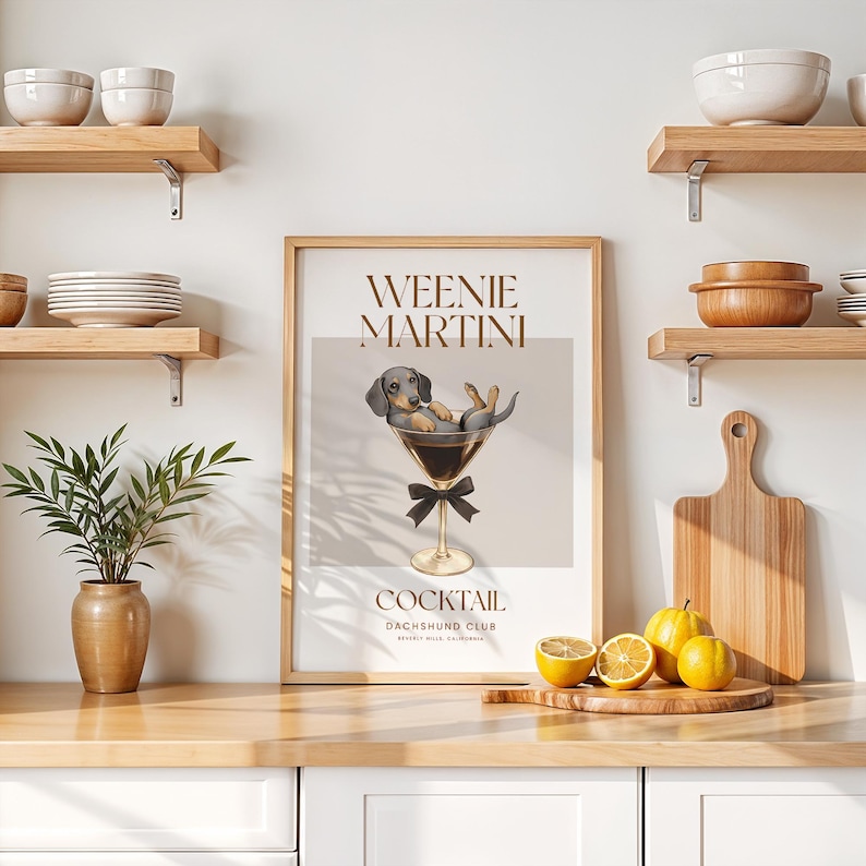 May include: A framed print featuring a dachshund dog relaxing in a martini glass. The print has the words "Weenie Martini" and "Cocktail Dachshund Club" in a stylish font. The print is displayed on a kitchen counter with lemons and a cutting board.