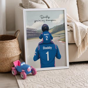 May include: A framed wall art print featuring a digital illustration of a child sitting on a parent's shoulders, both wearing blue racing suits. The child's suit says Matty 2 and the parent's says Daddy 1. Text reads: Daddy. you're my champion.