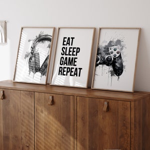 May include: Three framed prints with a gaming theme. The left print features a pair of black headphones with a watercolor effect. The center print has the text "Eat Sleep Game Repeat" in black. The right print shows a black video game controller with a watercolor effect.