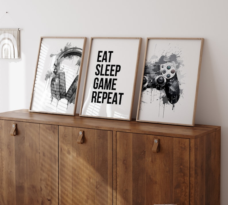 May include: Three framed prints with a gaming theme. The left print features a pair of black headphones with a watercolor effect. The center print has the text "Eat Sleep Game Repeat" in black. The right print shows a black video game controller with a watercolor effect.