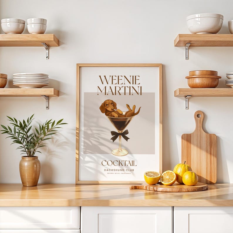 May include: A framed print featuring a dachshund dog relaxing in a martini glass, with the text "Weenie Martini Cocktail Dachshund Club". The print has a light brown background and a black bow. The image is displayed on a wooden countertop.