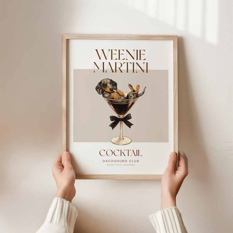 May include: A framed print featuring a dachshund dog relaxing in a martini glass. The print has the words "Weenie Martini" at the top and "Cocktail Dachshund Club" at the bottom. The dog is black, brown, and white.