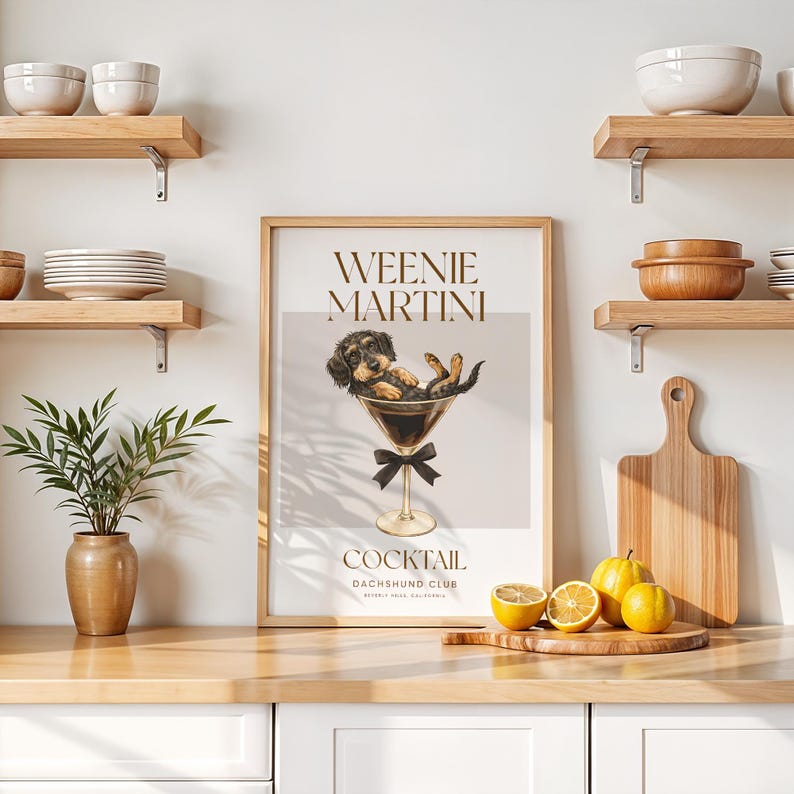 May include: A framed print featuring a dachshund dog relaxing in a martini glass. The print has the words "Weenie Martini" and "Cocktail Dachshund Club" in a stylish font. The image is set on a kitchen counter with lemons and a cutting board.