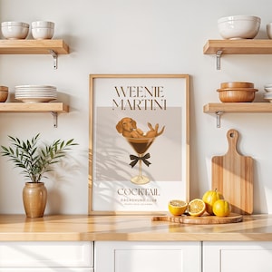 May include: A framed print with the words "Weenie Martini Cocktail Dachshund Club" features a dachshund dog relaxing in a martini glass. The print is in a light brown frame, with a black bow tie on the glass. Lemons and a cutting board are on the counter.
