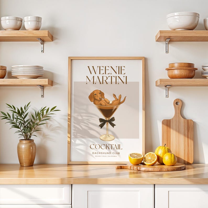 May include: A framed print with the words "Weenie Martini Cocktail Dachshund Club" features a dachshund dog relaxing in a martini glass. The print is in a light brown frame, with a black bow tie on the glass. Lemons and a cutting board are on the counter.